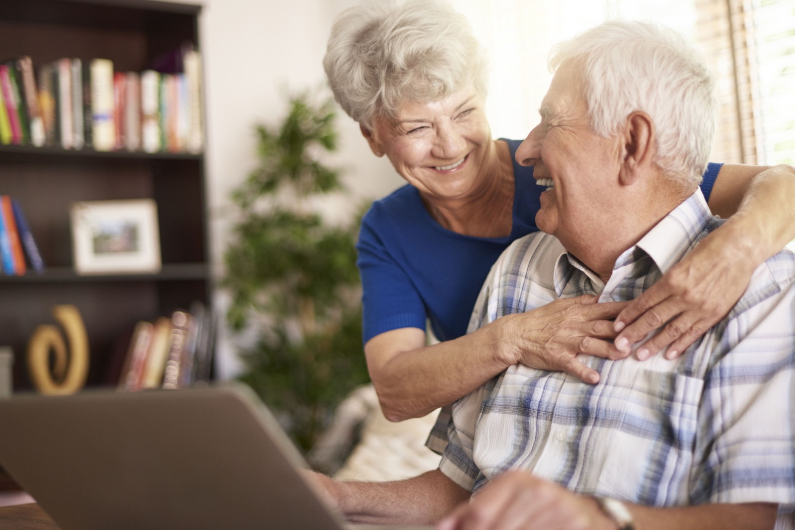 casal de idosos com homem no computador e mulher abraçando os ombros dele sorrindo felizes